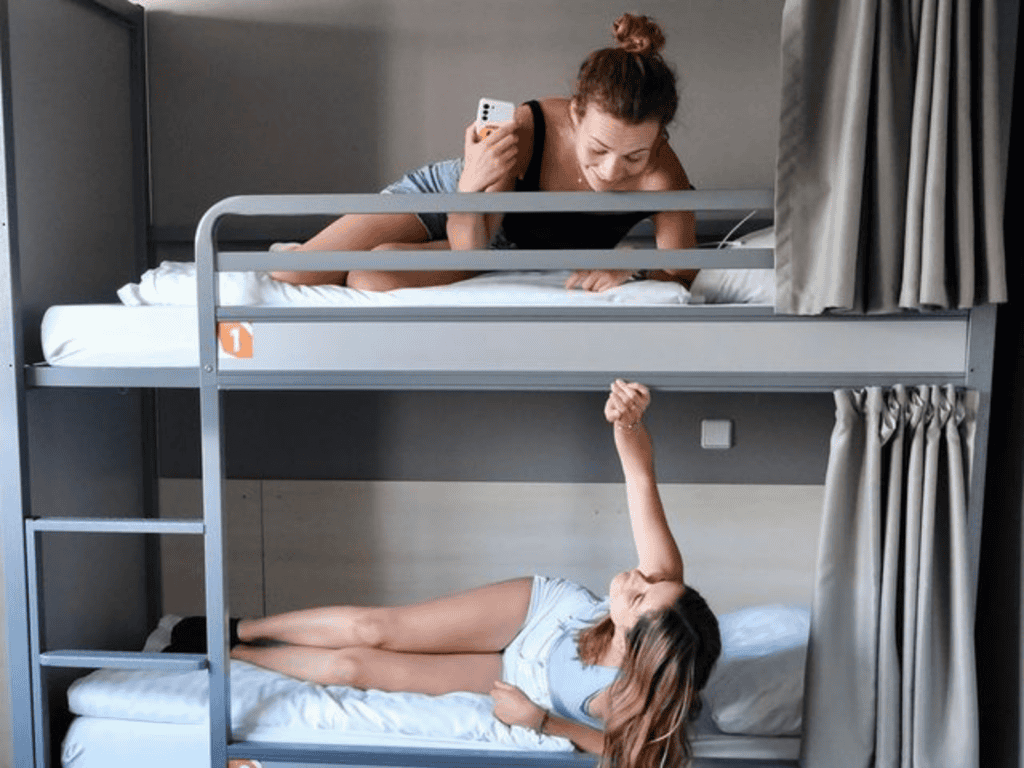 Young woman on top bunk talking on phone, girl on bottom bunk reaching up, hostel room scene.
