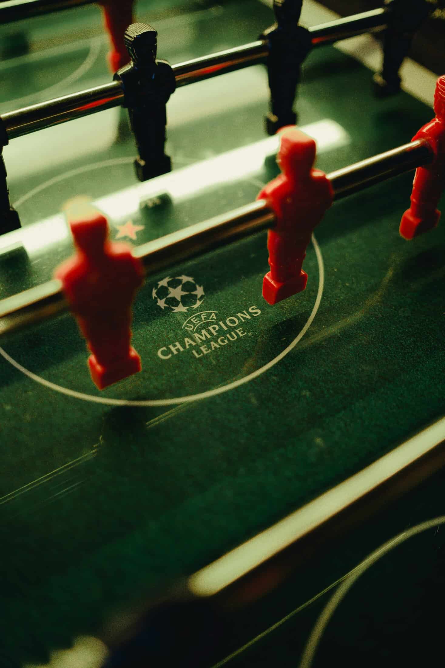 Foosball table with red and blue players, UEFA Champions League logo visible underneath, lively spor.
