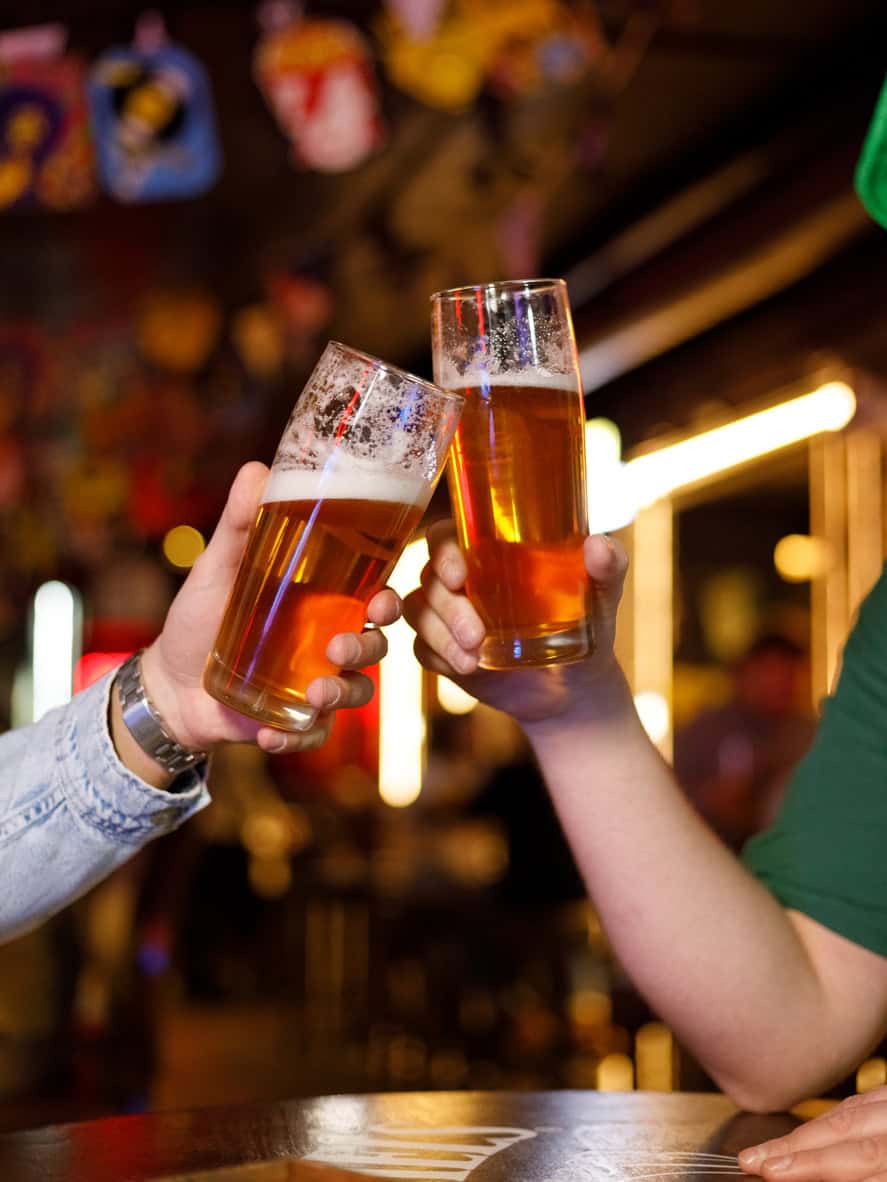 Two people clinking glasses of beer in a lively pub setting.