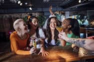 Group of diverse women celebrating with drinks at a bar, wearing a birthday crown.