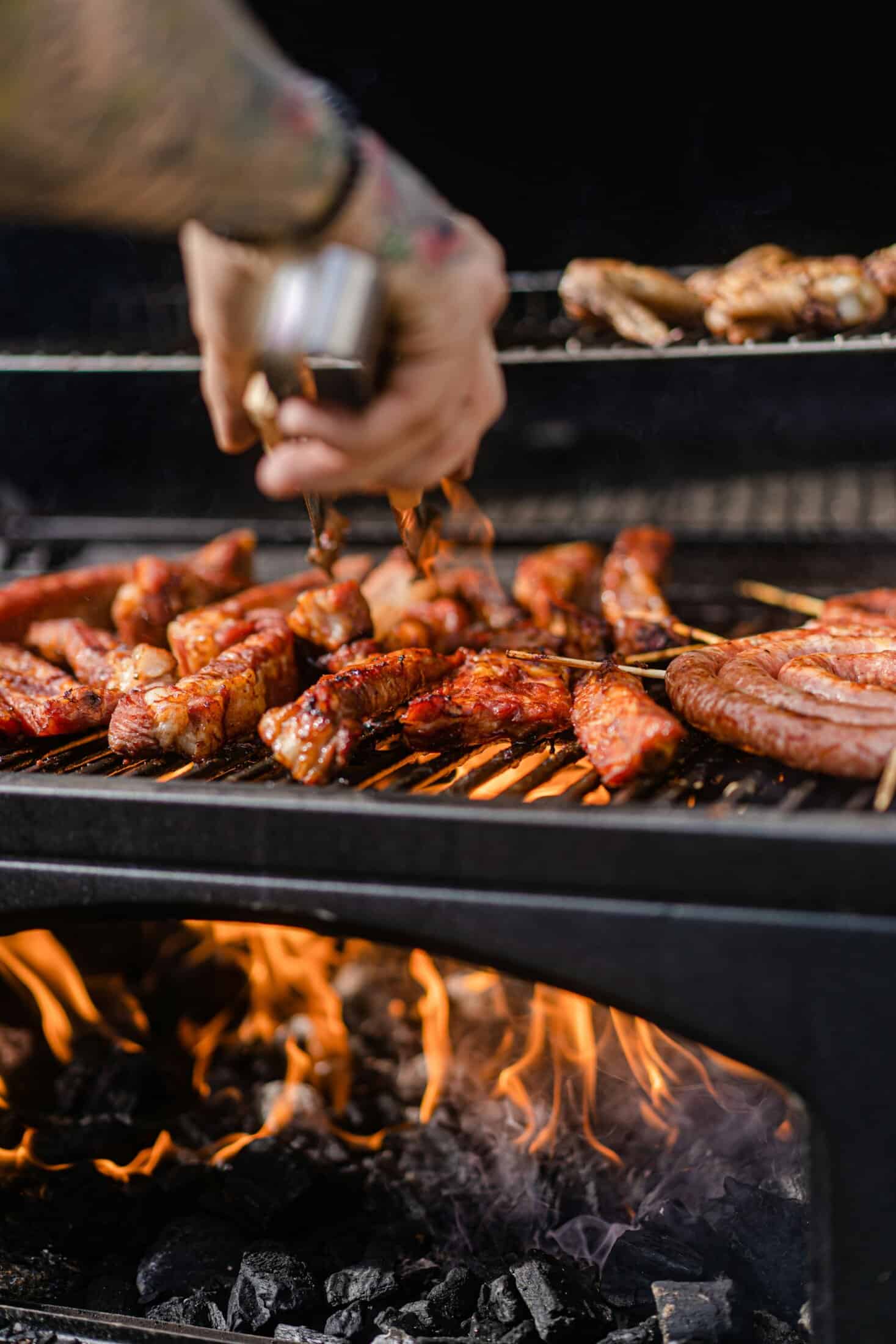 Grilling meat on barbecue with sauce being applied, outdoor cooking scene.